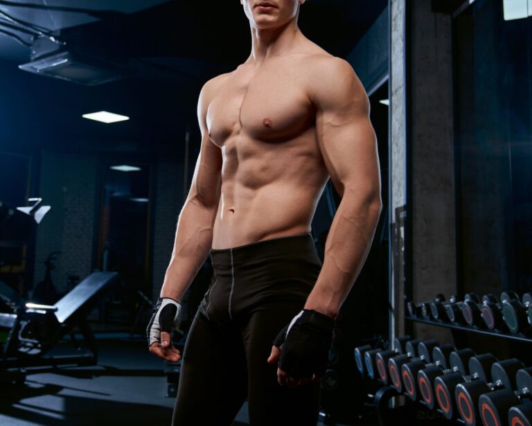 Side view of shirtless bodybuilder posing and looking at camera. Portrait of muscular handsome man with perfect body in gym in dark atmosphere. Concept of bodybuilding, healthy lifestyle.